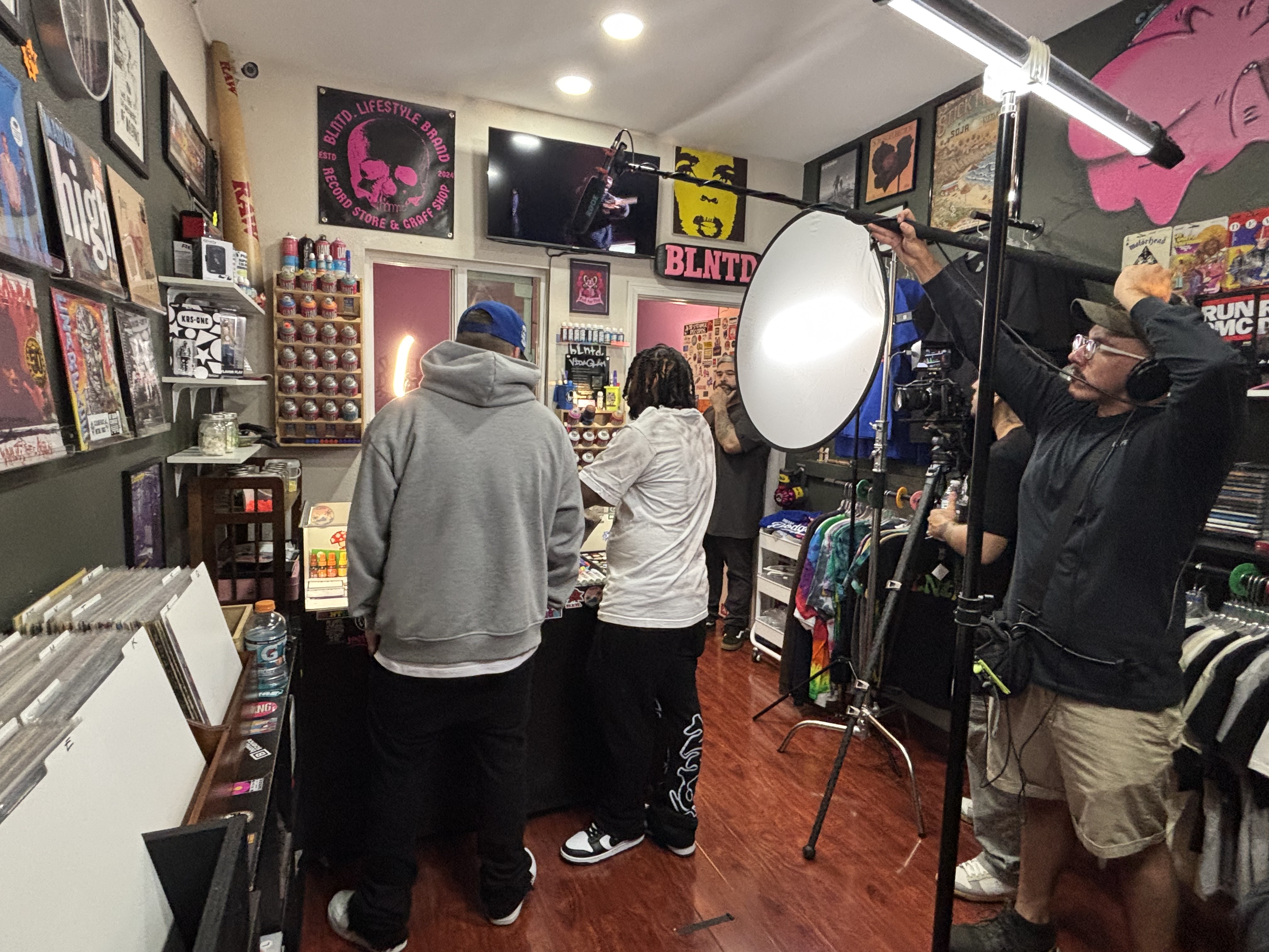 Behind the scenes of filming inside BLNTD., with cast, crew, lighting and sound equipment among clothing and record displays